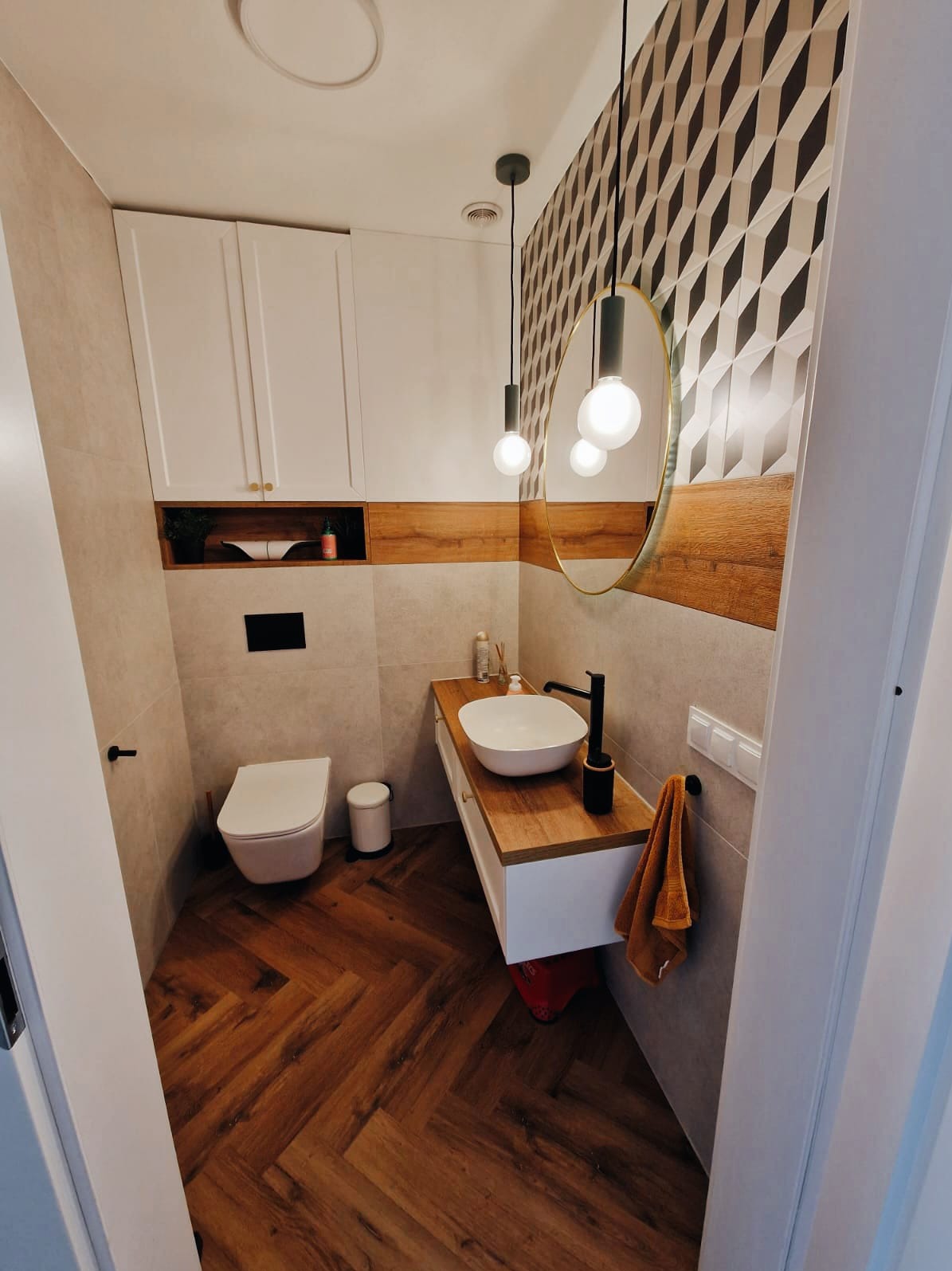 Nowoczesna łazienka z drewnianą podłogą ułożoną w jodełkę, białą umywalką nablatową na drewnianym blacie, wiszącymi szafkami i geometryczną tapetą w czarno-białe romby.