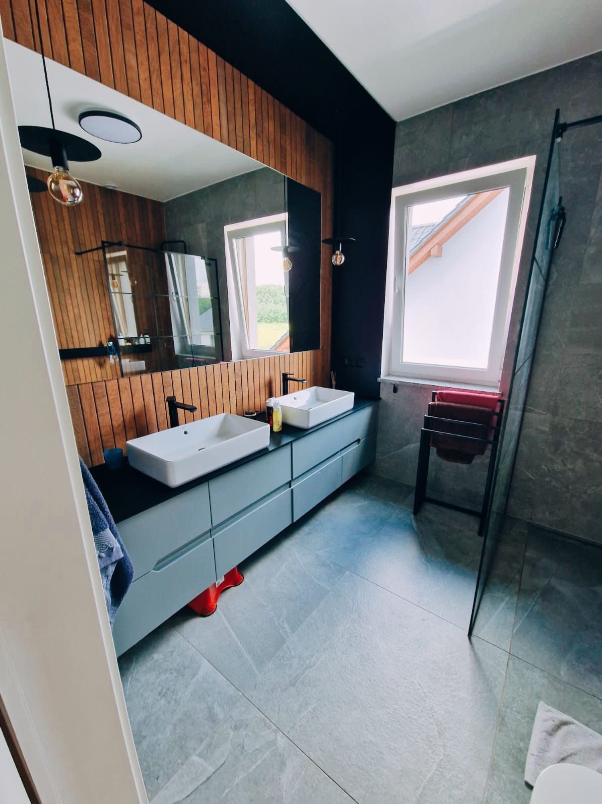 Nowoczesna łazienka z drewnianą ścianą, dwoma białymi umywalkami nablatowymi na szarej szafce, szarą podłogą z płytek i kabiną prysznicową typu walk-in z czarnymi elementami, widoczne czerwone...