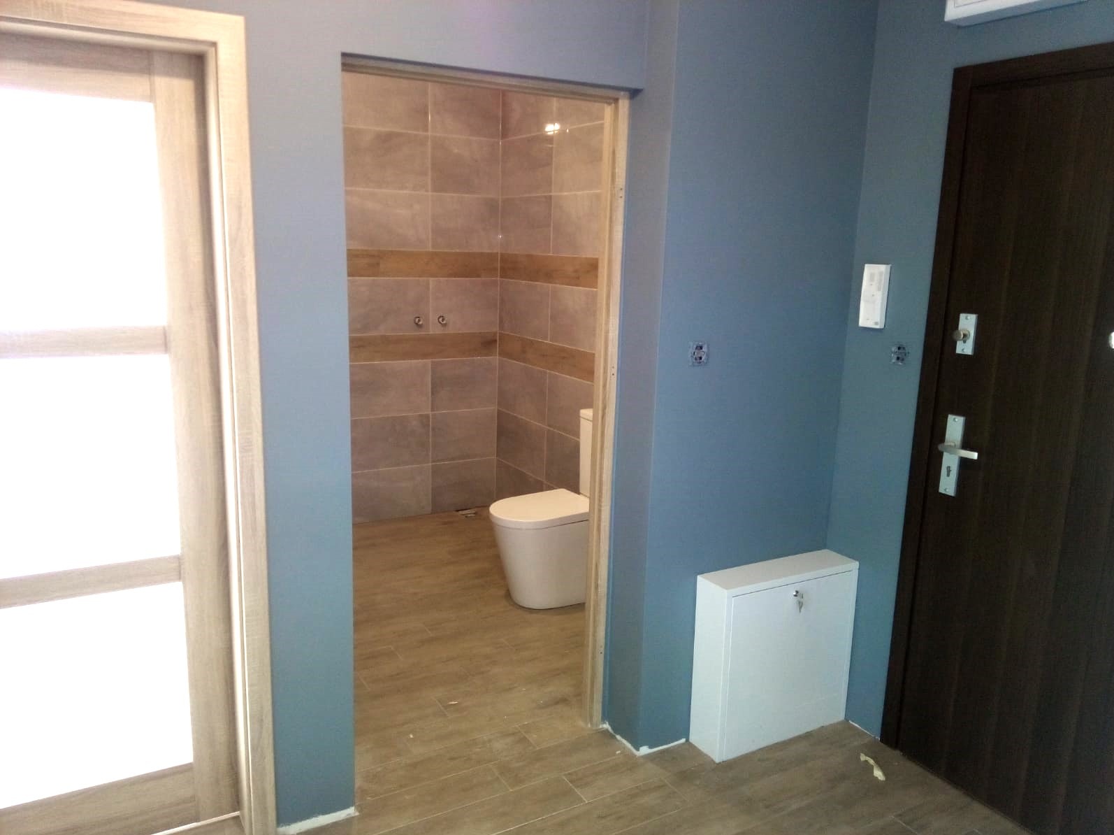 Widok na wykończoną łazienkę z toaletą, beżowe płytki na ścianach i podłodze, widoczne z korytarza z niebieskimi ścianami i brązowymi drzwiami wejściowymi.