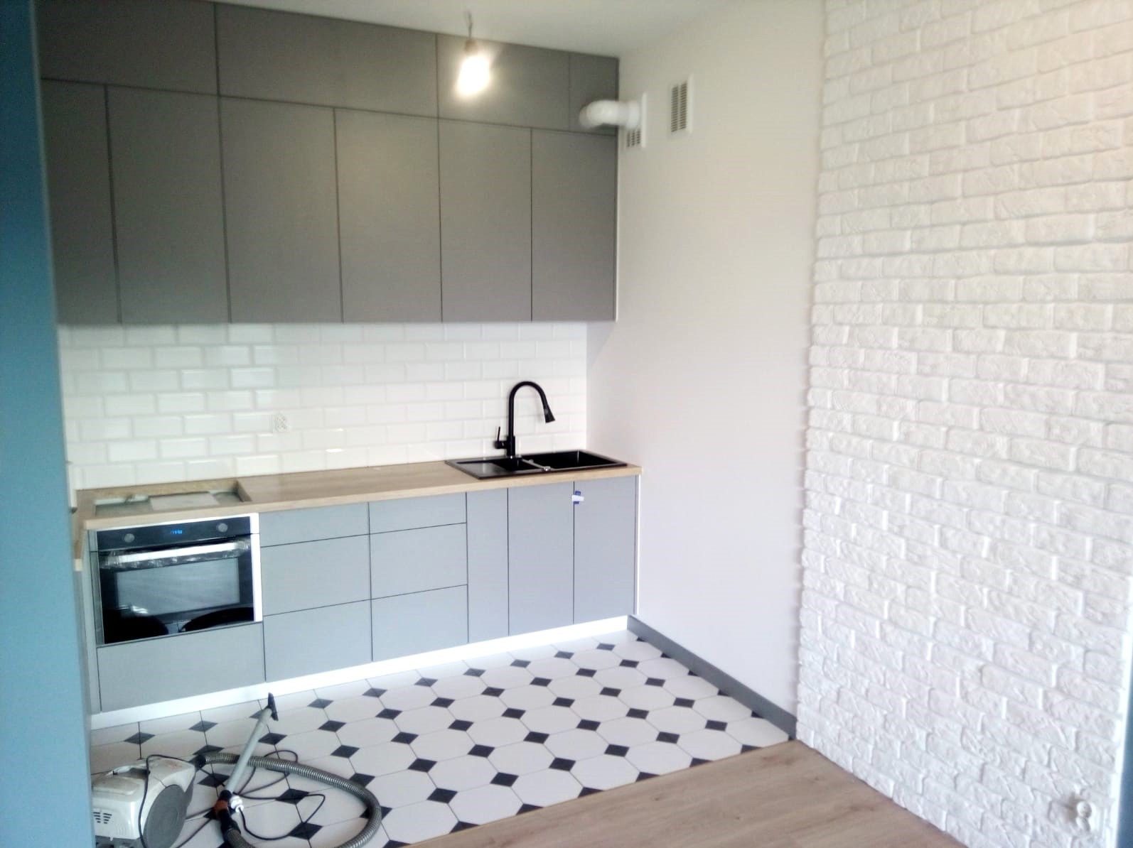 Szara zabudowa kuchenna z białymi płytkami metra nad blatem, czarnym zlewem i baterią, oraz białą ścianą z cegły dekoracyjnej. Na podłodze czarno-biała mozaika, widoczny odkurzacz.