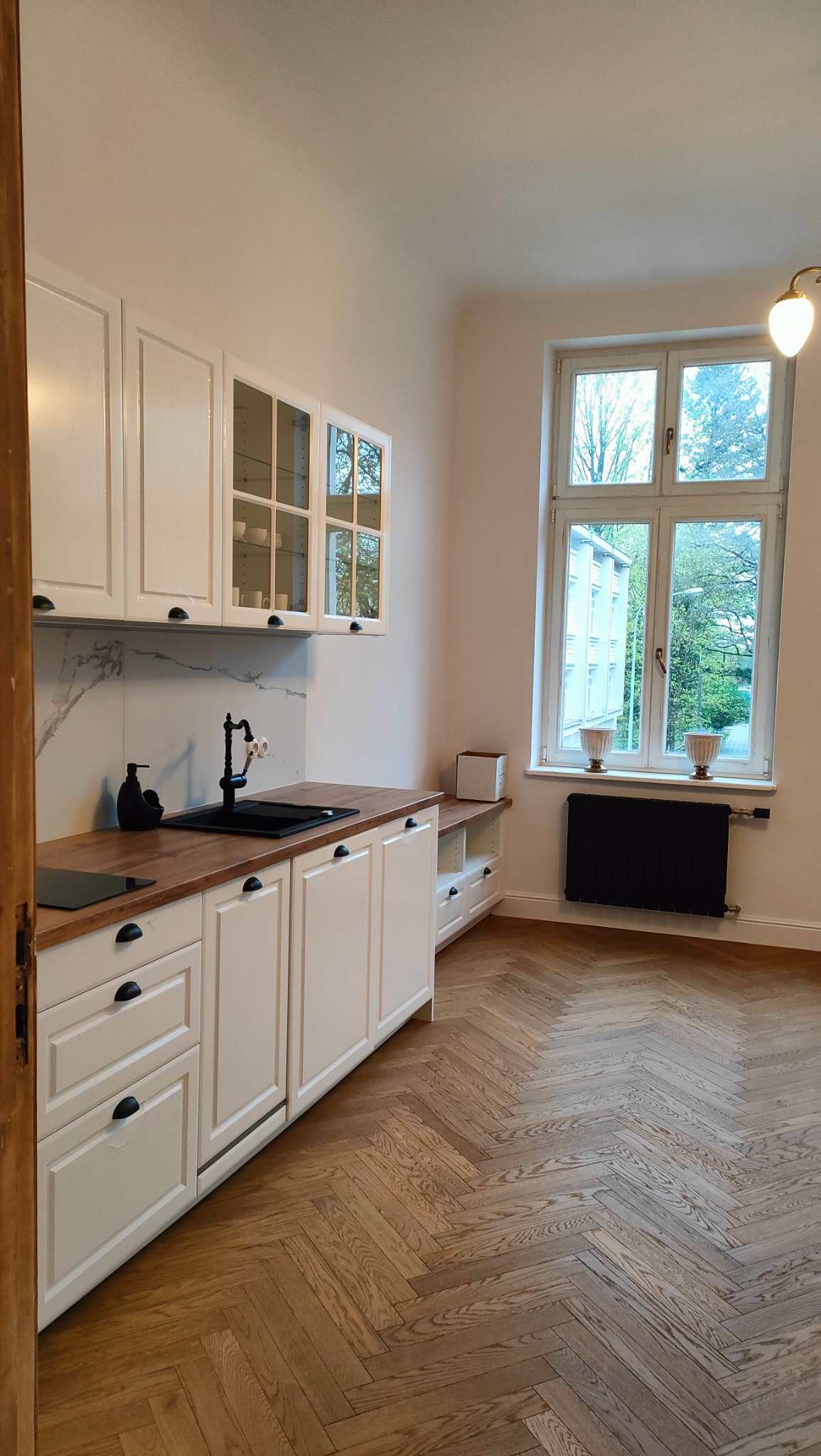 Jasna kuchnia z białymi szafkami, drewnianym blatem i czarną armaturą, parkiet w jodełkę, widok na zieleń za oknem.