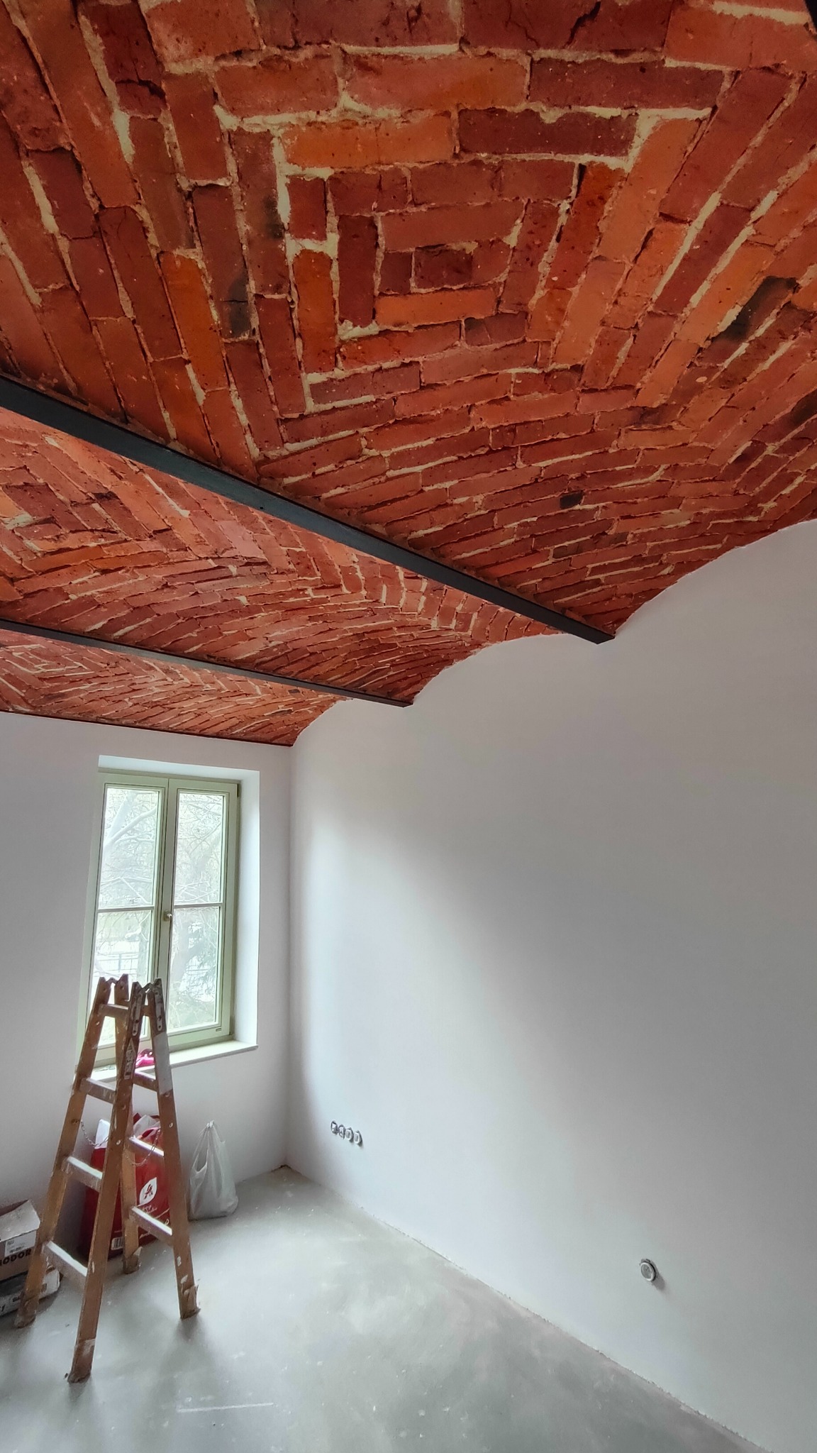 Ceglany sufit łukowy z metalowymi belkami, widok z dołu, drabina oparta o ścianę, okno z widokiem na drzewa bez liści, surowy beton na podłodze, ściany pomalowane na biało.