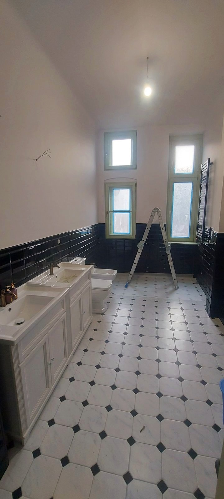Wnętrze łazienki w trakcie remontu: biała umywalka z szafką, bidet, czarne płytki na ścianach, biało-czarna podłoga, drabina oparta o ścianę z oknami z zielonymi ramami, widoczne przewody elektryczne.