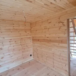 Wnętrze domu w trakcie wykańczania, ściany, podłoga i sufit wyłożone jasnym drewnem, widoczne kable elektryczne wystające z sufitu, brak listew przypodłogowych.
