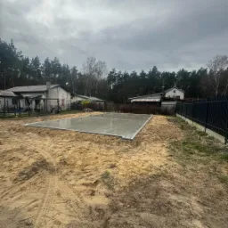 Wylana płyta fundamentowa na piaszczystym terenie, widoczne ślady opon, w tle domy i las pod pochmurnym niebem.