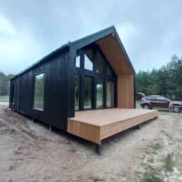 Domy z drewna Glinka - Nowoczesny, czarny dom z drewna na podwyższeniu, z dużymi oknami i drewnianym tarasem, w tle las i samochód z otwartym bagażnikiem.