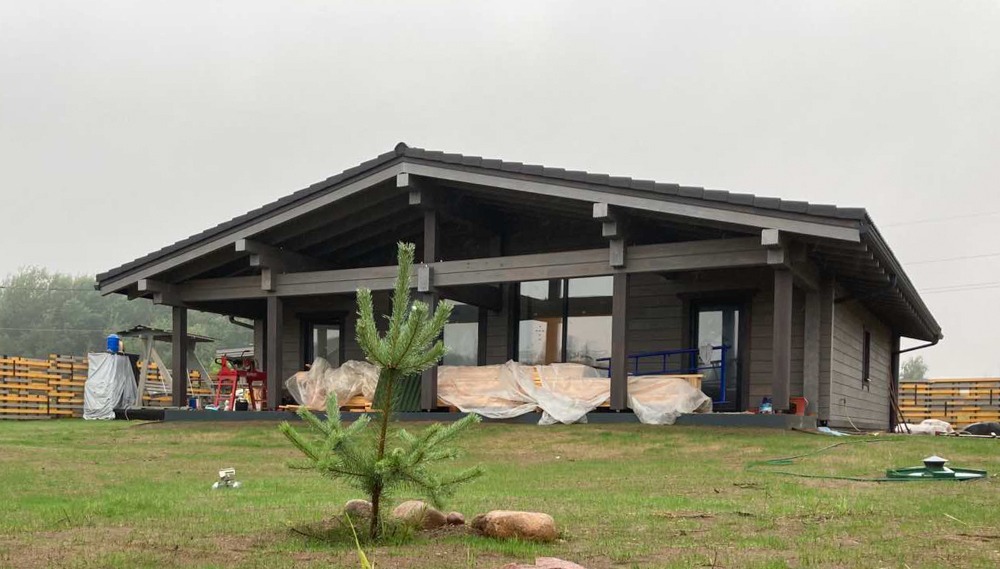 Nowoczesny parterowy dom z bali w trakcie budowy, widoczne materiały budowlane zabezpieczone folią, na pierwszym planie mała sosna.