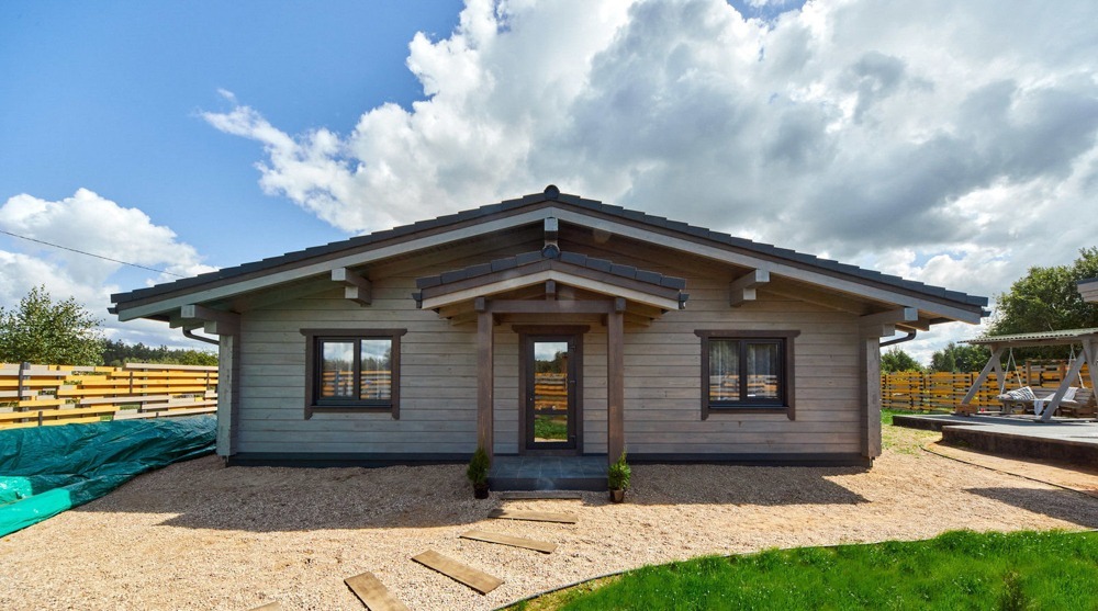 Parterowy dom z bali w odcieniach szarości, z ciemnobrązową stolarką okienną i drzwiową, otoczony żwirowym podłożem i elementami drewnianego chodnika, widok z przodu w słoneczny dzień.