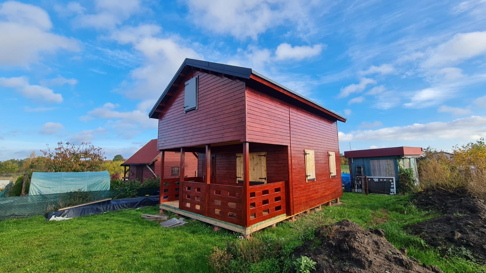 Czerwony dom szkieletowy z drewnianymi okiennicami, widoczny z zewnątrz na tle błękitnego nieba z chmurami, z widocznymi elementami konstrukcyjnymi u podstawy.
