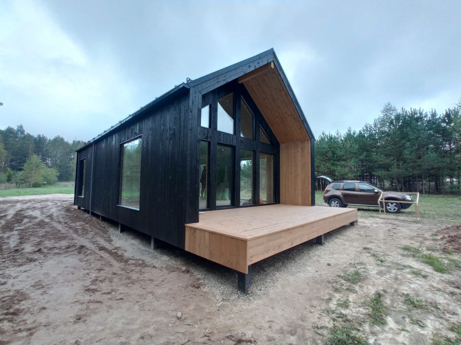 Nowoczesny, czarny dom z drewna na podwyższeniu, z dużymi oknami i drewnianym tarasem, w tle las i samochód z otwartym bagażnikiem.