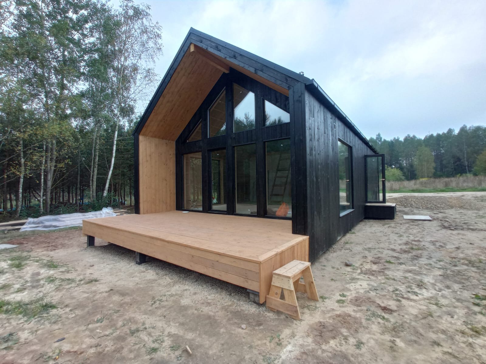 Nowoczesny, czarny dom z drewna z dużymi oknami, z surowym wykończeniem elewacji i drewnianym tarasem, w otoczeniu drzew i niezagospodarowanego terenu.