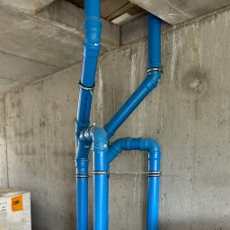 Serhii Kalchuk HIPPO - Kompleksowy układ niebieskich rur kanalizacyjnych z metalowymi obejmami, zamontowany przy surowym betonowym suficie i ścianie w nowym budynku w stanie deweloperskim. W tle widoczne opakowania...