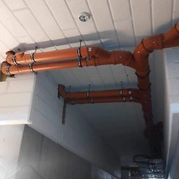 Serhii Kalchuk HIPPO - Pomarańczowe rury kanalizacyjne zamontowane pod sufitem z betonowych bloczków, widoczne metalowe obejmy mocujące i fragment instalacji elektrycznej.