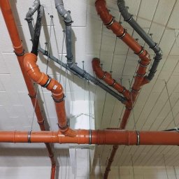 Serhii Kalchuk HIPPO - Pomarańczowe i szare rury kanalizacyjne podwieszone do sufitu z betonu komórkowego, widoczne metalowe obejmy i pręty podtrzymujące, instalacja w surowym stanie.
