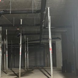 Serhii Kalchuk HIPPO - Surowy strop z widocznymi czarnymi rurami kanalizacyjnymi podwieszonymi na metalowych wspornikach, liczne stemple budowlane podtrzymujące konstrukcję, mokra betonowa podłoga w szarym pomieszczeniu.