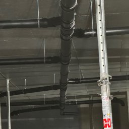 Serhii Kalchuk HIPPO - Widok z dołu na szare rury kanalizacyjne pod sufitem, podparte metalowymi stemplami budowlanymi.