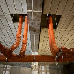 Serhii Kalchuk HIPPO - Pomarańczowe rury kanalizacyjne podwieszone pod stropem z płyt OSB, widoczne uchwyty mocujące i etykiety na rurach, szare ściany w tle.