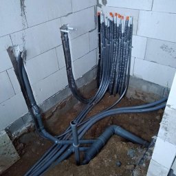 Serhii Kalchuk HIPPO - Surowe ściany z betonu komórkowego i świeżo ułożone rury kanalizacyjne z szarego PCV w wykopie, widoczne także pionowe rury instalacji elektrycznej w peszlach z pomarańczowymi zabezpieczeniami...