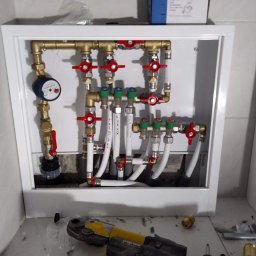 Serhii Kalchuk HIPPO - Rozdzielacz instalacji grzewczej w białej szafce, widoczny wodomierz, zawory z czerwonymi i zielonymi pokrętłami oraz białe rury. Na podłodze narzędzia: zaciskarka i wiertarko-wkrętarka.