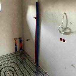 Serhii Kalchuk HIPPO - Rozłożona instalacja ogrzewania podłogowego na metalowej siatce w surowym pomieszczeniu, widoczne rury doprowadzające i przygotowane otwory w ścianie.