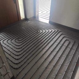 Serhii Kalchuk HIPPO - Rury ogrzewania podłogowego ułożone spiralnie na izolacji, widok z góry przed wylewką, krawędź ściany z widoczną listwą przypodłogową.