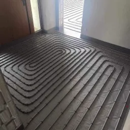 Rury ogrzewania podłogowego ułożone spiralnie na izolacji, widok z góry przed wylewką, krawędź ściany z widoczną listwą przypodłogową.