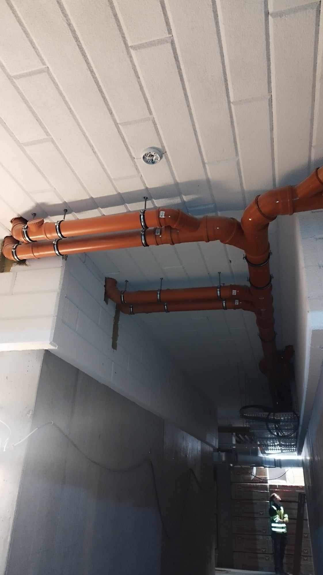 Pomarańczowe rury kanalizacyjne zamontowane pod sufitem z betonowych bloczków, widoczne metalowe obejmy mocujące i fragment instalacji elektrycznej.