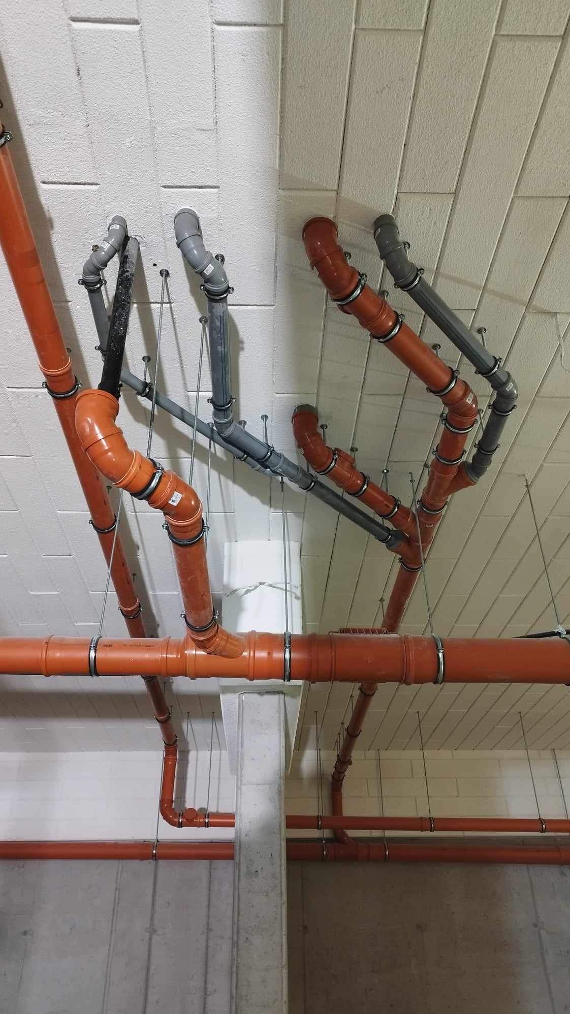 Pomarańczowe i szare rury kanalizacyjne podwieszone do sufitu z betonu komórkowego, widoczne metalowe obejmy i pręty podtrzymujące, instalacja w surowym stanie.