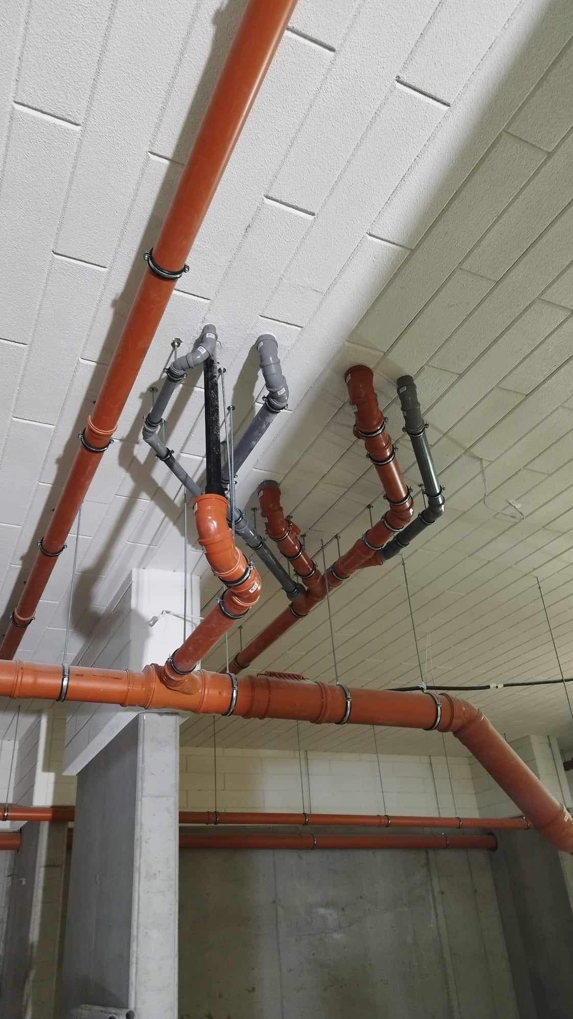 Pomarańczowe i szare rury kanalizacyjne zamontowane pod sufitem z betonowych płyt, widoczne stalowe obejmy mocujące i fragment ściany z betonu.