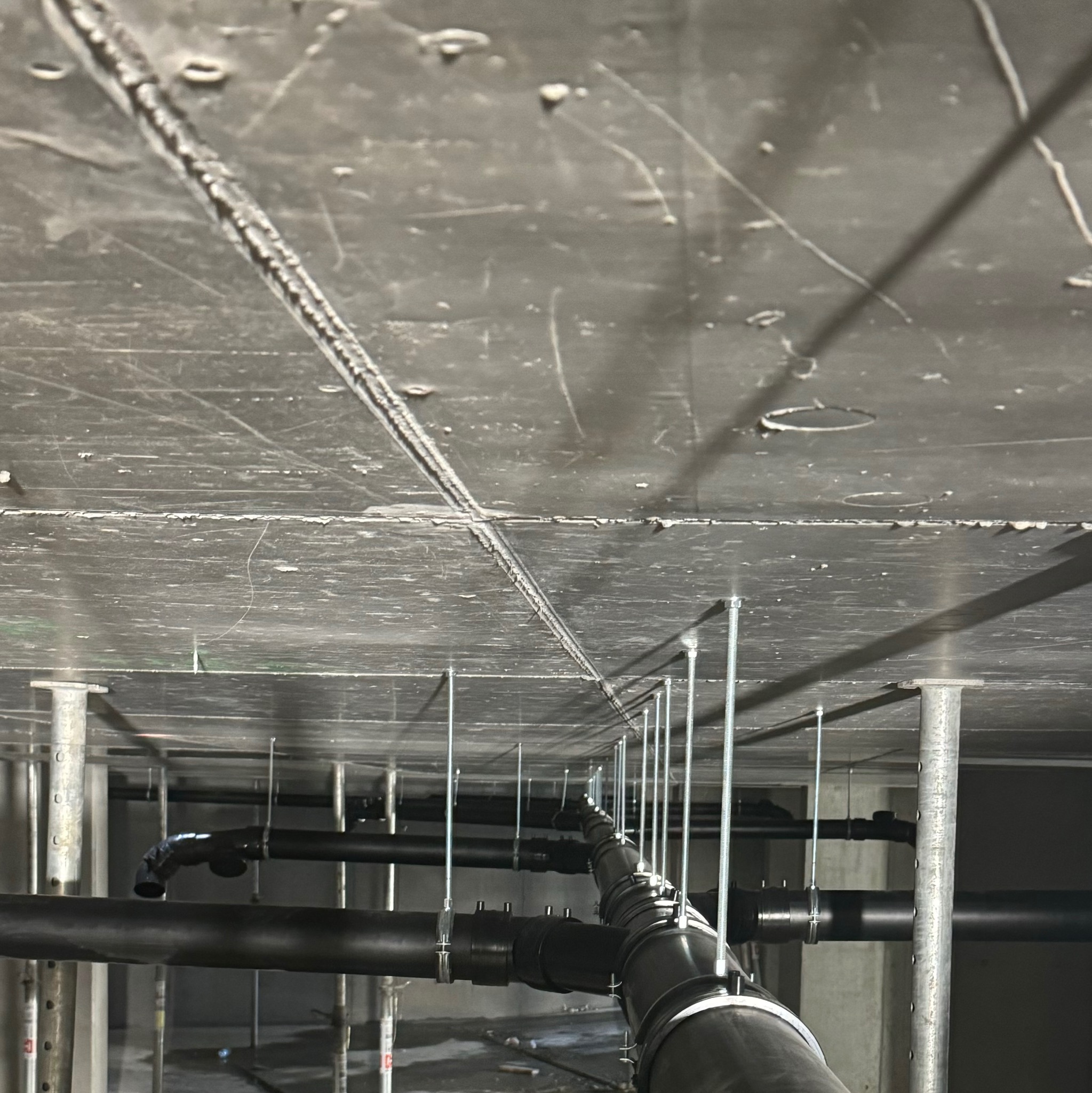 Czarne rury kanalizacyjne podwieszone do sufitu za pomocą metalowych prętów, widoczne z perspektywy podłogi w surowym wnętrzu budynku.