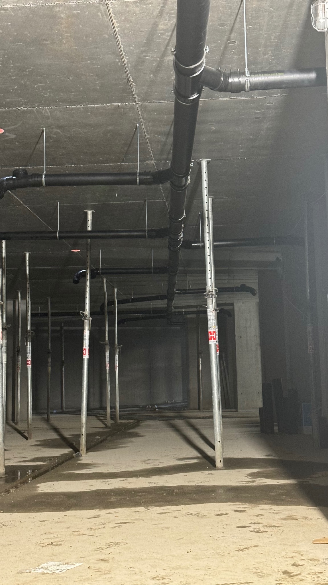 Surowy strop z widocznymi czarnymi rurami kanalizacyjnymi podwieszonymi na metalowych wspornikach, liczne stemple budowlane podtrzymujące konstrukcję, mokra betonowa podłoga w szarym pomieszczeniu.