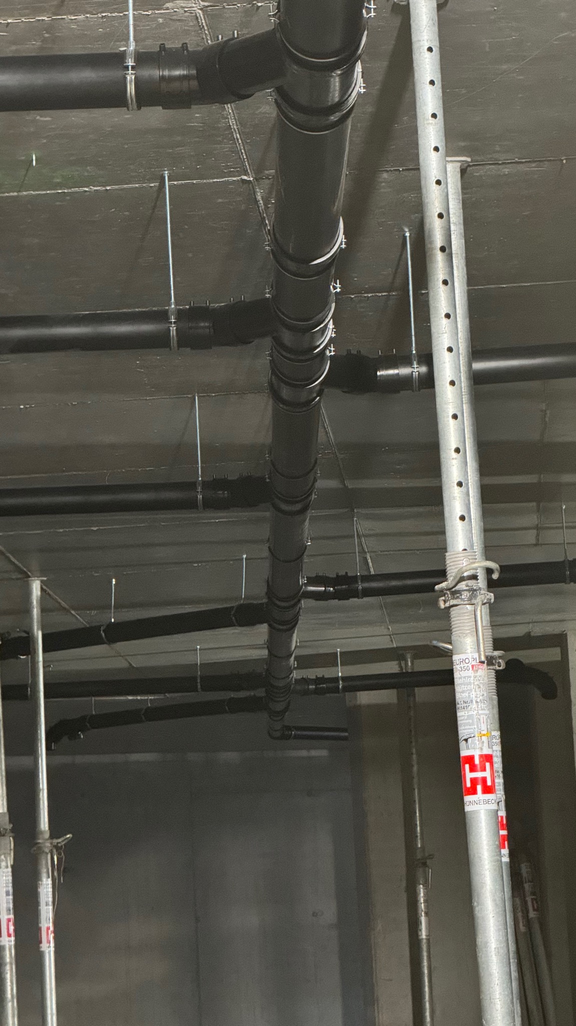 Widok z dołu na szare rury kanalizacyjne pod sufitem, podparte metalowymi stemplami budowlanymi.