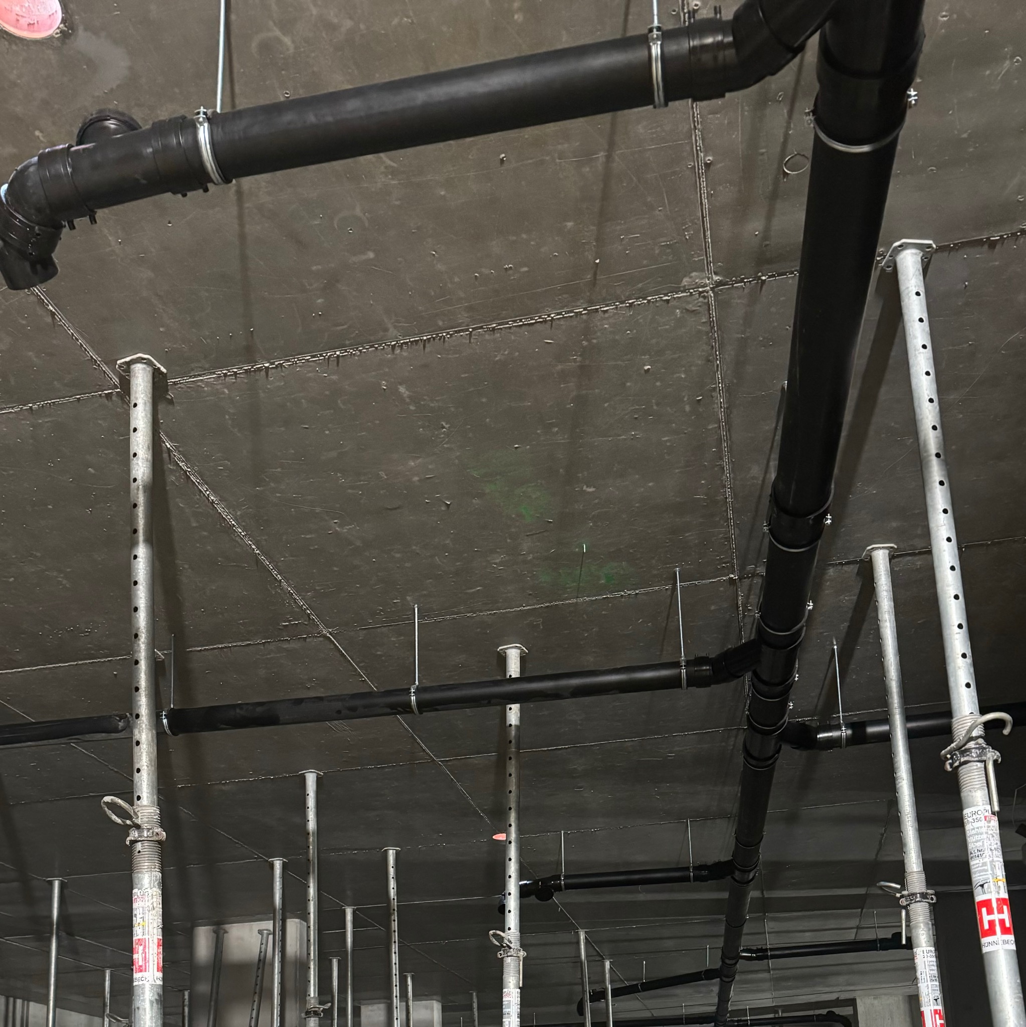 Widok z dołu na sufit w trakcie budowy, z widocznymi czarnymi rurami kanalizacyjnymi podwieszonymi za pomocą metalowych obejm i wspartych na regulowanych metalowych podporach budowlanych.