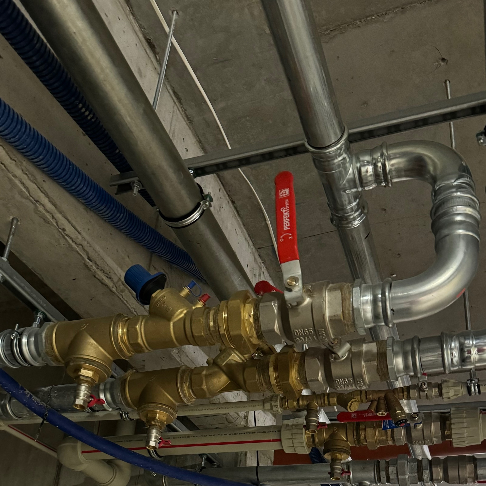 Skomplikowany system rur i zaworów z mosiądzu i stali pod surowym betonowym stropem, widoczne niebieskie rury karbowane.