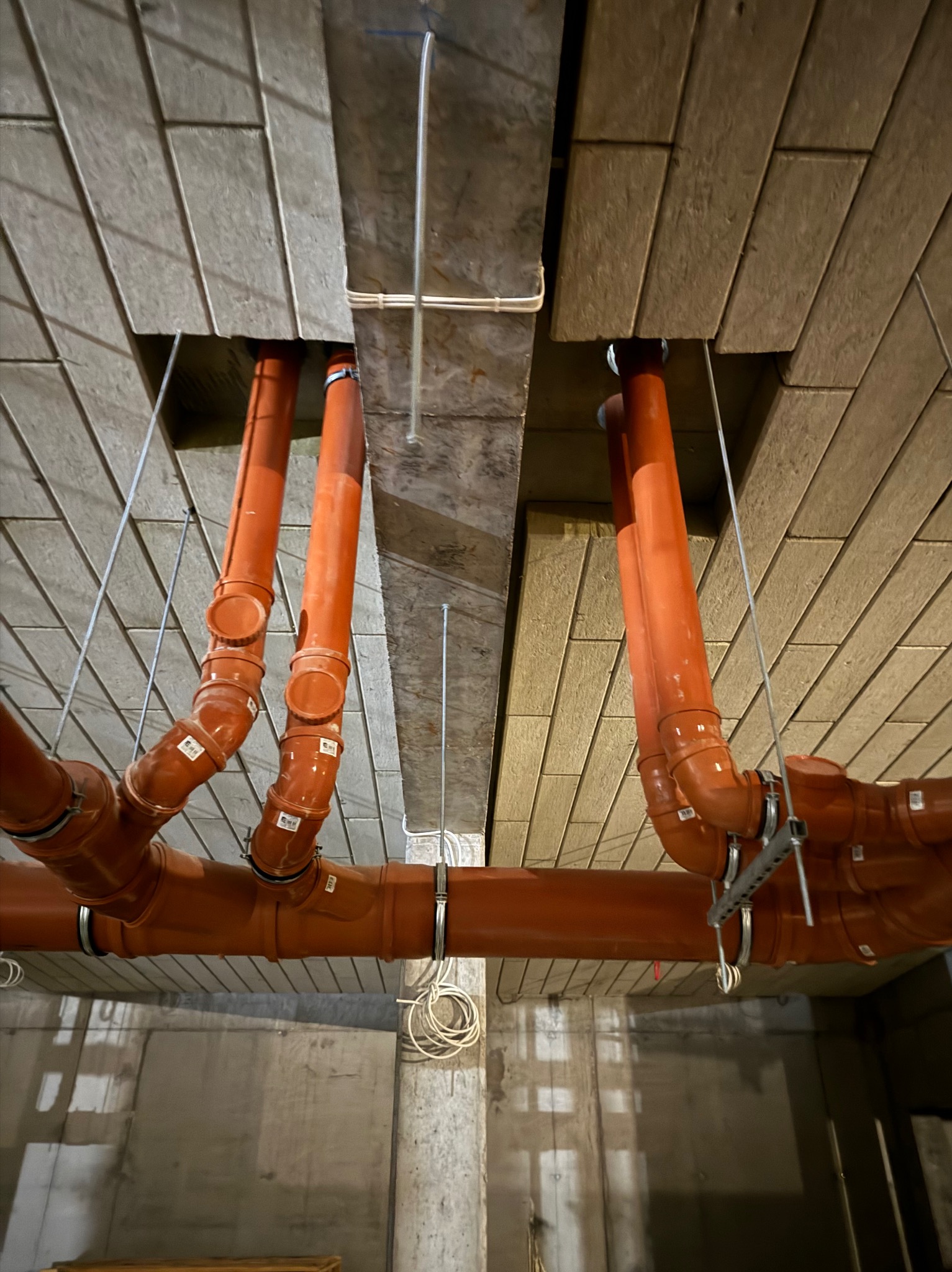Pomarańczowe rury kanalizacyjne podwieszone pod stropem z płyt OSB, widoczne uchwyty mocujące i etykiety na rurach, szare ściany w tle.