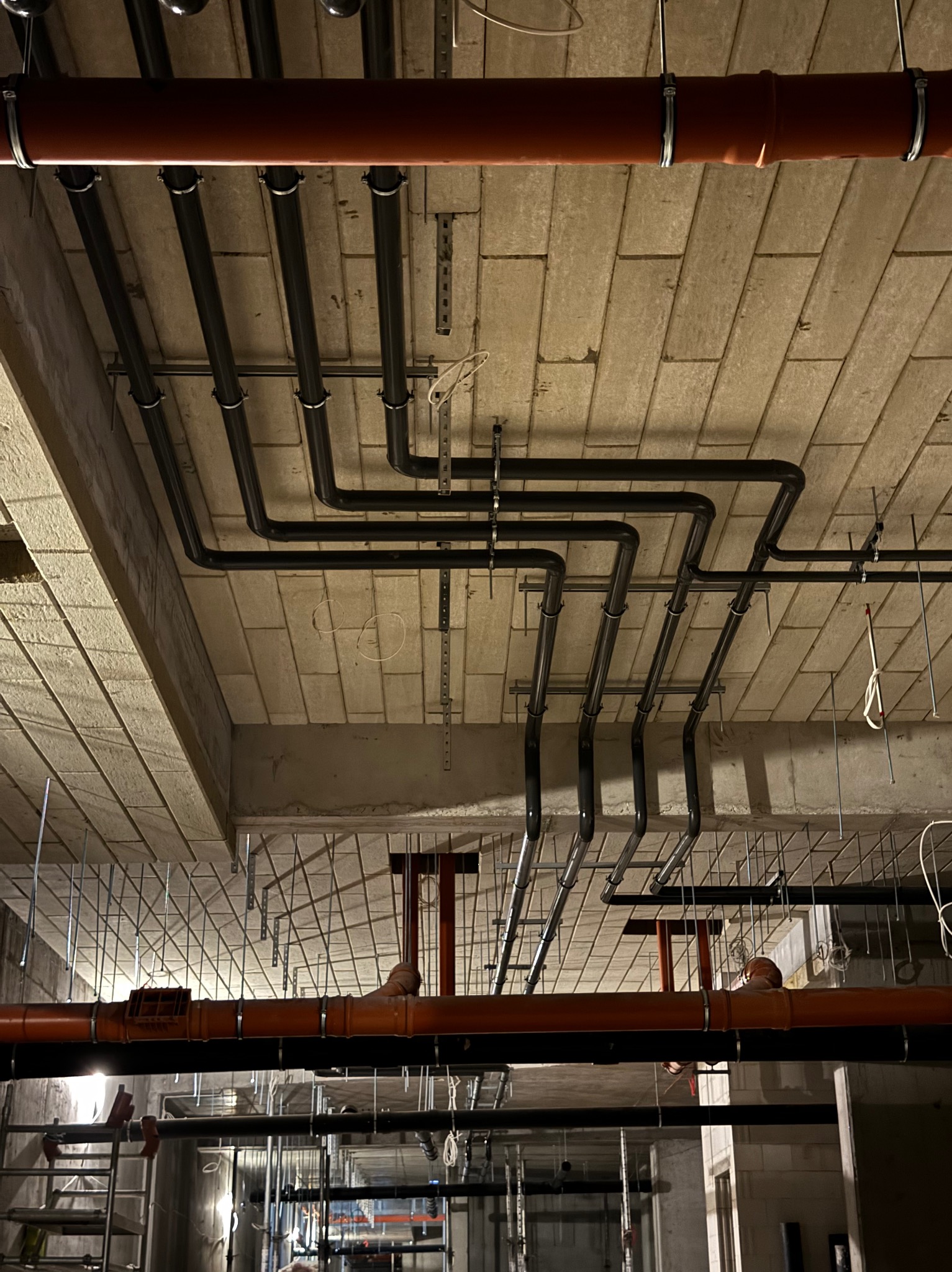 Sufit z widocznymi rzędami rur w kolorze czarnym i pomarańczowym, zabezpieczonych metalowymi obejmami, na tle surowego betonowego stropu z widoczną fakturą bloczków.