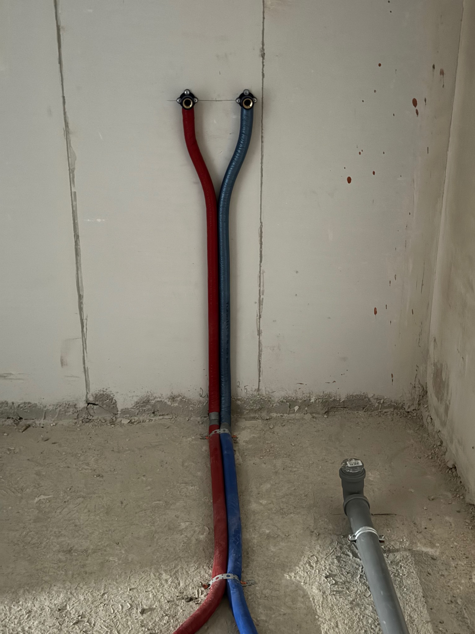 Czerwona i niebieska rura doprowadzająca wodę, zamocowane do surowej ściany w trakcie prac instalacyjnych, widoczny odpływ kanalizacyjny w podłodze.