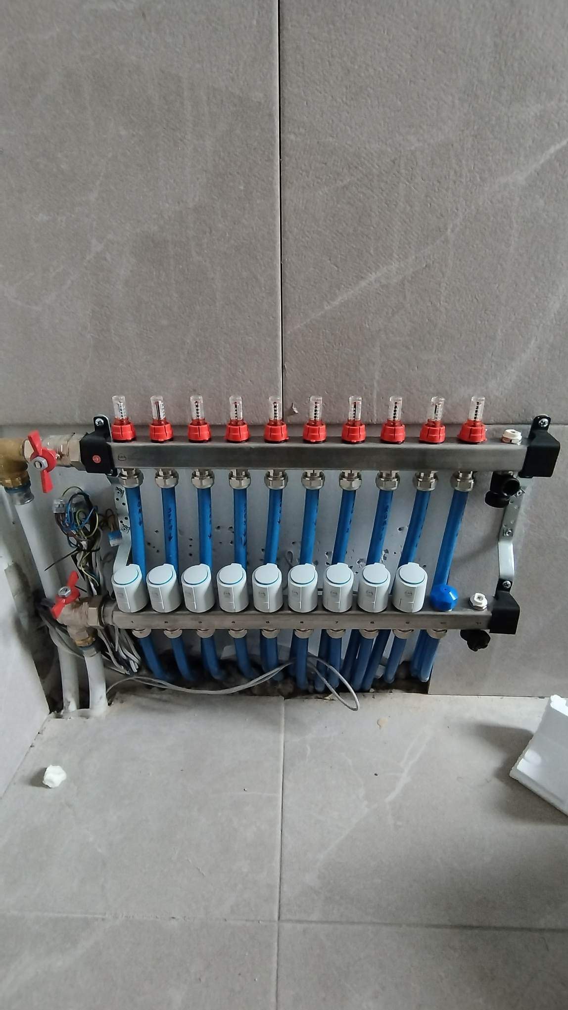 Rozdzielacz ogrzewania podłogowego z niebieskimi rurami i białymi zaworami termostatycznymi, zamontowany na szarej ścianie z płytek, widoczne okablowanie i elementy instalacji.