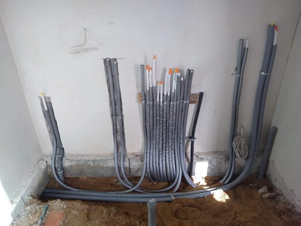 Wewnętrzna instalacja hydrauliczna w trakcie realizacji: rury w izolacji prowadzone w ścianie i pod podłogą, z widocznymi mocowaniami i fragmentem wykopu w ziemi.