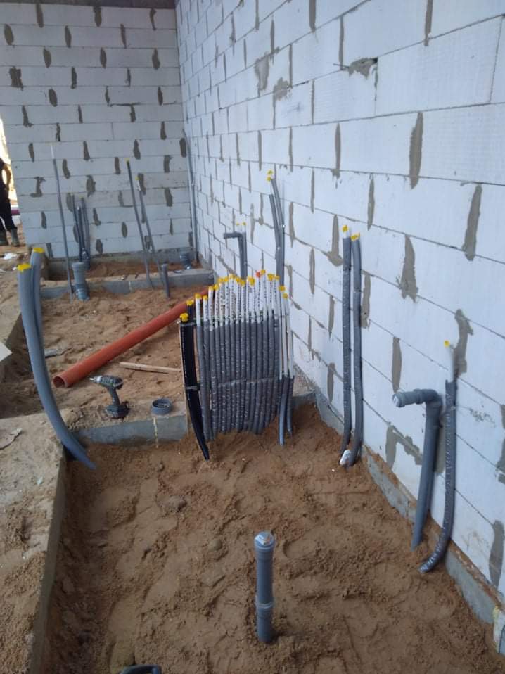 Wykop z piaskiem i fragmentem ściany z betonu komórkowego, liczne rury w izolacji z żółtymi zaślepkami, narzędzia w tle, etap prac instalacyjnych.