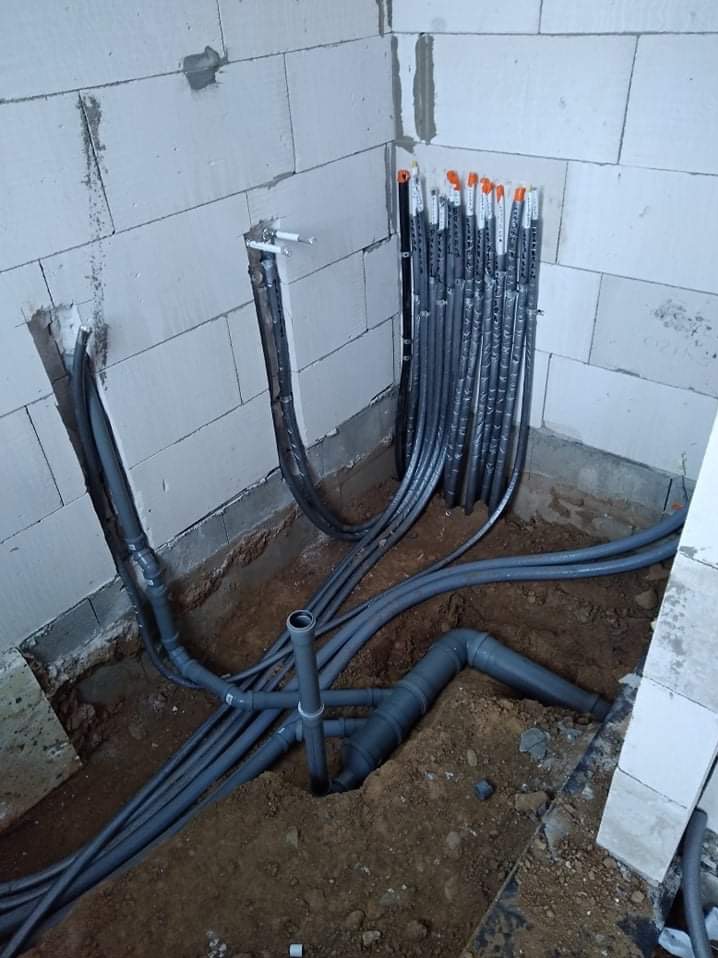 Surowe ściany z betonu komórkowego i świeżo ułożone rury kanalizacyjne z szarego PCV w wykopie, widoczne także pionowe rury instalacji elektrycznej w peszlach z pomarańczowymi zabezpieczeniami...