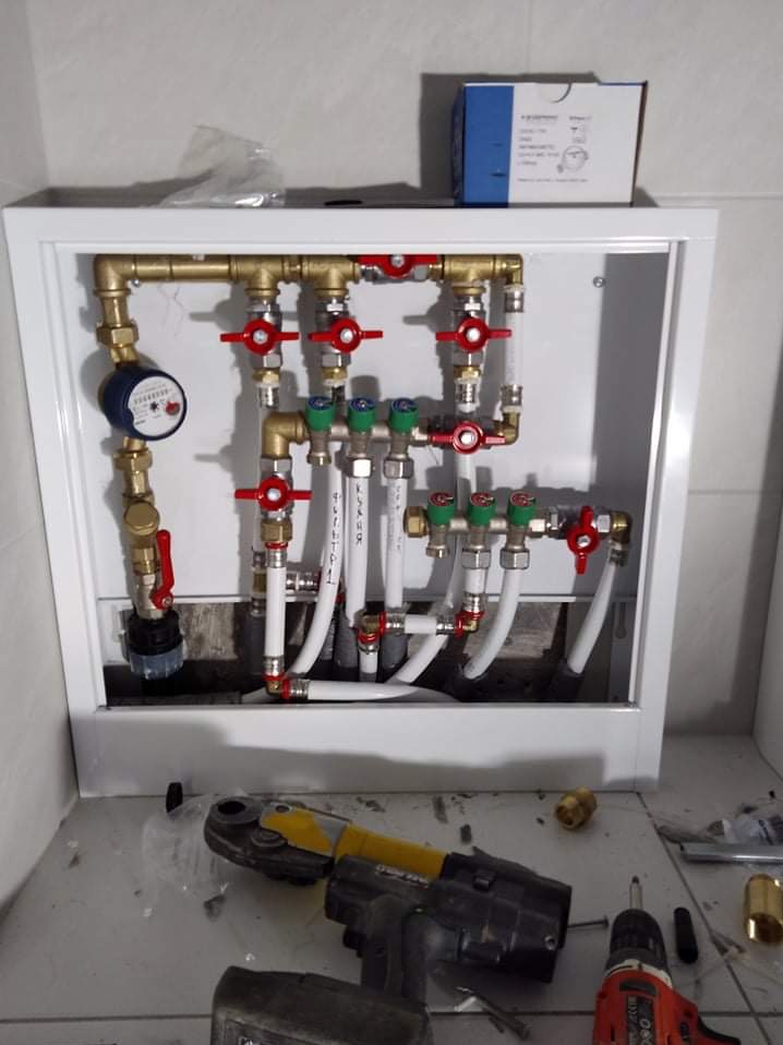 Rozdzielacz instalacji grzewczej w białej szafce, widoczny wodomierz, zawory z czerwonymi i zielonymi pokrętłami oraz białe rury. Na podłodze narzędzia: zaciskarka i wiertarko-wkrętarka.