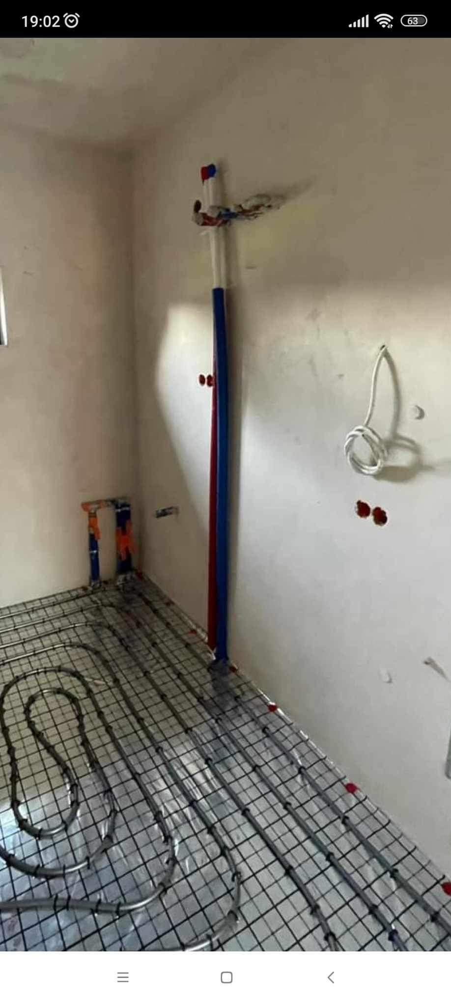 Rozłożona instalacja ogrzewania podłogowego na metalowej siatce w surowym pomieszczeniu, widoczne rury doprowadzające i przygotowane otwory w ścianie.