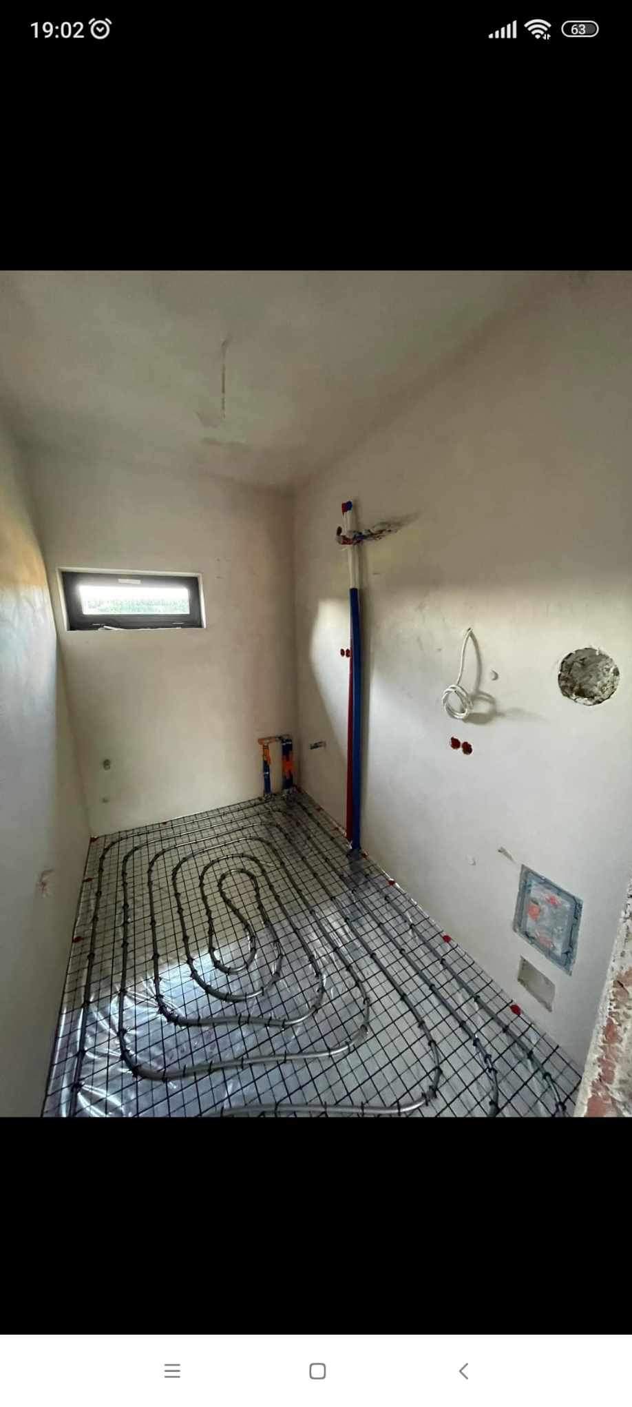 Wnętrze łazienki w trakcie instalacji ogrzewania podłogowego: rury grzewcze ułożone na siatce, widoczne pionowe rury doprowadzające wodę, surowe ściany z widocznymi śladami prac instalacyjnych...