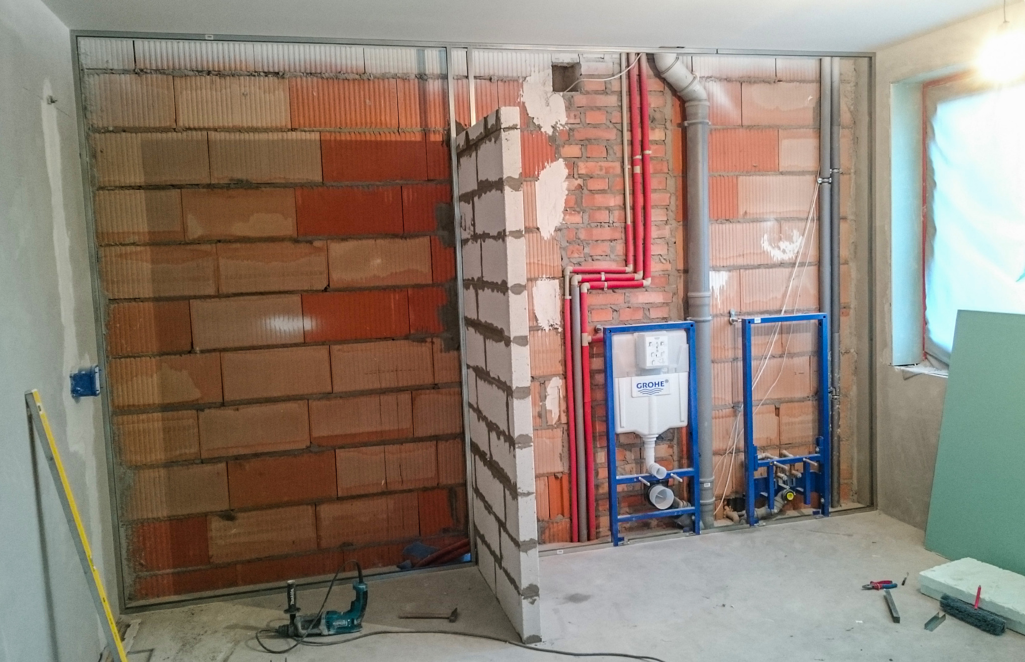 Wnętrze łazienki w trakcie remontu: widoczne ceglane ściany z metalową konstrukcją pod zabudowę, instalacja sanitarna z niebieską ramą i białą podtynkową spłuczką Grohe, czerwone rury instalacji...