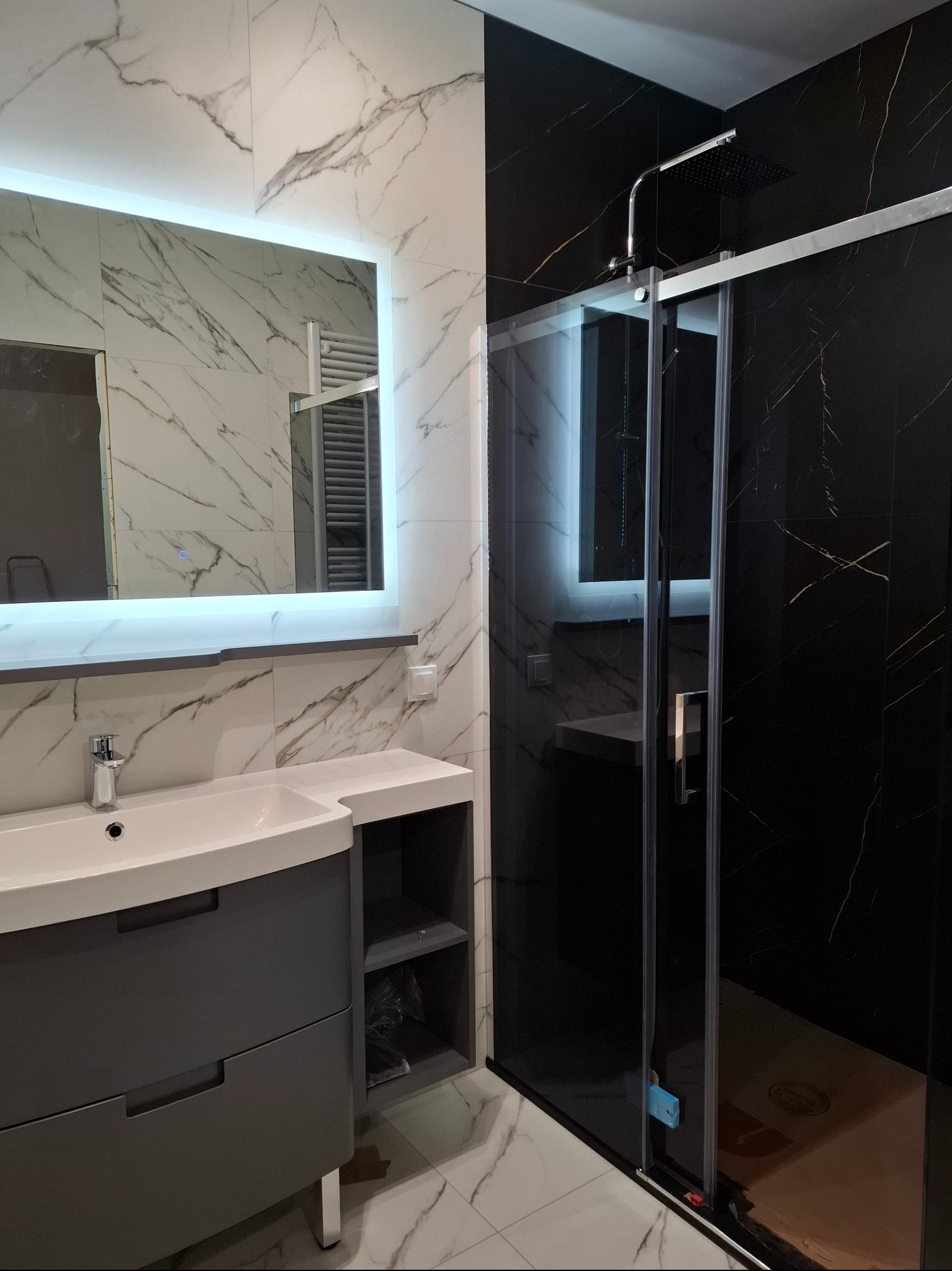 Nowoczesna łazienka z czarno-białymi płytkami imitującymi marmur, podświetlanym lustrem, szarą szafką pod umywalką i kabiną prysznicową ze szklanymi przesuwnymi drzwiami.