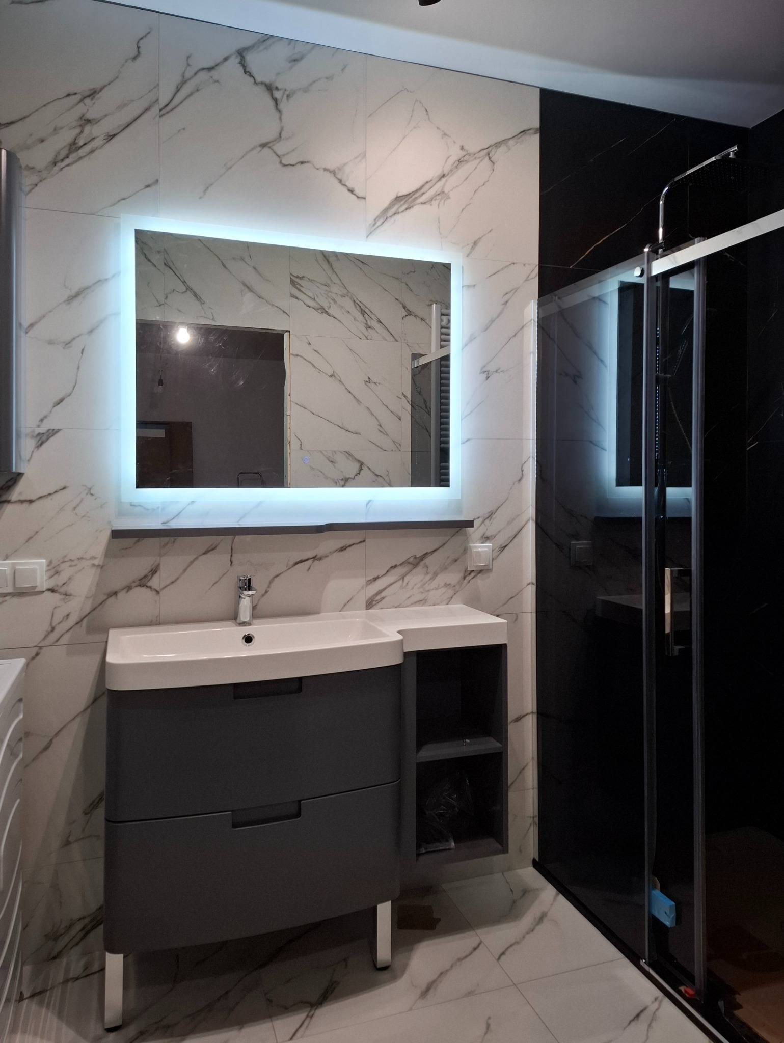 Nowoczesna łazienka z umywalką w szarej szafce, podświetlanym lustrem i kabiną prysznicową z czarnymi elementami, wykończona płytkami imitującymi marmur.