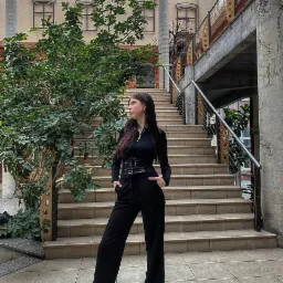 Kobieta w czarnym stroju pozuje na tle schodów w przestronnym wnętrzu z roślinami i przeszklonym dachem, z ręką w kieszeni i wzrokiem skierowanym w bok.