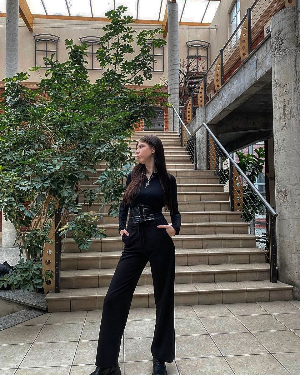 Kobieta w czarnym stroju pozuje na tle schodów w przestronnym wnętrzu z roślinami i przeszklonym dachem, z ręką w kieszeni i wzrokiem skierowanym w bok.