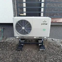 Biała jednostka zewnętrzna pompy ciepła Galmet Prima R32 zamontowana na stalowej konstrukcji na żwirowym podłożu, widoczna łopata i wąż ogrodowy, montaż w toku.